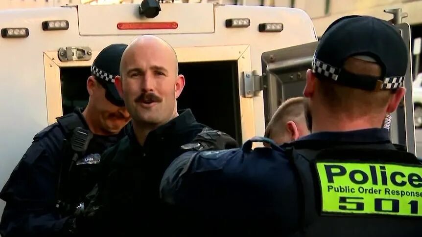 A bald man with a brown moustache looks at the camera while being handcuffed by police.